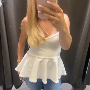 Vit peplum linnetopp med v-ringning - Säljer en vit peplum linnetopp med smala axelband och v-ringning. Toppen har en figurnära passform upptill och en utsvängd volang nertill som ger en snygg siluett. Perfekt att styla med jeans eller kjol för en fräsch look.