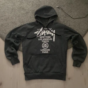 Grå Stussy hoodie  - Säljer en svart hoodie från Stussy med vit logga och tryck på bröstet med städer som New York, Los Angeles, Tokyo, London och Paris. Klassisk känguruficka och huva med snörning. Perfekt för dig som gillar streetwear och vill sticka ut.