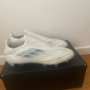 Adidas F50 vita fotbollsskor - Säljer ett par Adidas F50 fotbollsskor i vitt med blå detaljer. Skorna har en strömlinjeformad design utan snörning, med mönstrad ovandel och genomskinlig sula med blå och gula dobbar. Perfekta för dig som vill sticka ut på planen.