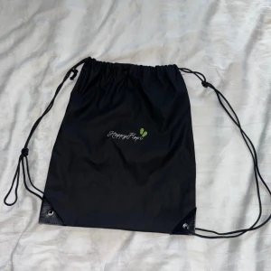 Svart gympåse från HappyFlops - En svart gympåse med dragsko och logga från HappyFlops framtill. Väskan har tunna snören som axelband och förstärkta hörn nertill. Perfekt för att bära träningskläder eller skor. Enkel och stilren design med liten grön detalj vid loggan.