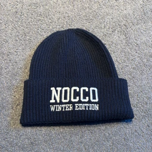 Mörkblå NOCCO mössa Winter Edition - Ribbstickad mörkblå mössa från NOCCO med uppvikt kant och broderad vit text 'NOCCO WINTER EDITION' framtill. Perfekt för kalla dagar och snygg att bära till vinterjackan. Denna går ej att köpa eftersom den ingick i nocco kalendern julen 2023 så finns bara ett par limiterade mössor av denna sort!