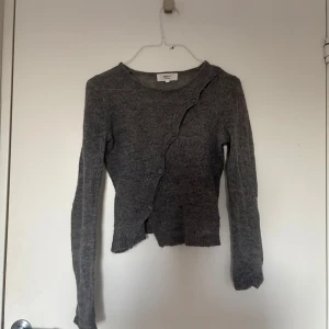 Grå kofta från Monki med asymmetrisk knäppning - Grå stickad kofta från Monki med lång ärm och unik asymmetrisk knäppning framtill. Koftan har ribbade muddar och en lätt croppad passform som ger en cool och avslappnad vibe. Perfekt för lager-på-lager under kyliga dagar.