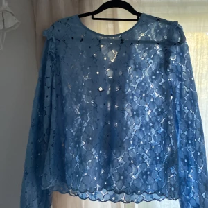 Blå spetsblus med snörning - Supersöt blå blus i spets med blommigt mönster och långa ärmar. Baksidan har snygg snörningsdetalj som ger en unik look. Perfekt för dig som gillar romantiska vibes och vill sticka ut lite extra. Blusen är lätt transparent och har rund halsringning.