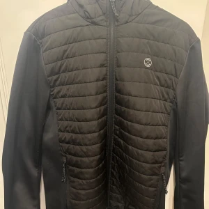 Svart jack and Jones jacka med huva - Snygg svart jacka med quiltad design och huva. Jackan har dragkedja framtill och ett stilrent märke på bröstet.