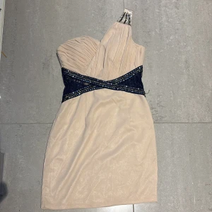 Champagnefärgad one shoulder-klänning - Snygg kortklänning från Little Mistress i champagnefärg med marinblå spets och paljettdetaljer i midjan. Klänningen har one shoulder-design med veckad överdel och glittriga detaljer vid axeln. Perfekt för dig som vill sticka ut med en elegant look.