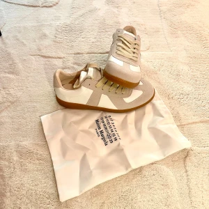 Maison Margiela sneakers beige/vit - Snygga sneakers från Maison Margiela i beige och vitt med klassisk gummisula. Skorna har paneler i mocka och skinn, samt beige snörning. Perfekta för dig som gillar stilrena och trendiga sneakers med exklusiv känsla.