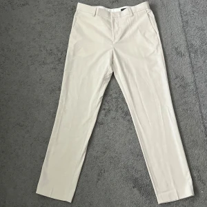 Beiga kostymbyxor slim fit från H&M - Snygga beiga kostymbyxor från H&M i slim fit-modell. Byxorna har klassisk design med hällor för bälte, snedställda fickor fram och passpoalfickor bak. Materialet är en mix av polyester, viskos och elastan för en skön känsla och bra passform.