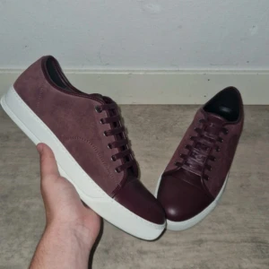 Vinröda sneakers från Lanvin - Säljer ett par stilrena vinröda sneakers från Lanvin med ovandel i mocka och detaljer i skinn. Skorna har vita platta sulor och klassisk snörning. Perfekta för dig som vill ha en clean och lyxig look med en twist av färg.