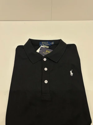 Svart pikétröja från Polo Ralph Lauren - Klassisk svart pikétröja från Polo Ralph Lauren med vit broderad logga på bröstet och tre vita knappar framtill. Tröjan har krage och är tillverkad i ett mjukt bomullsmaterial som känns skönt mot huden. Skriv för annan info