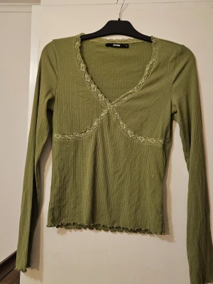 Grön topp med spetsdetaljer - Snygg olivgrön långärmad topp från Bikbok med ribbad struktur och dekorativa spetsdetaljer längs v-ringningen och över bysten. Toppen har en feminin passform och vågig kant nertill, perfekt för att lyfta din outfit.