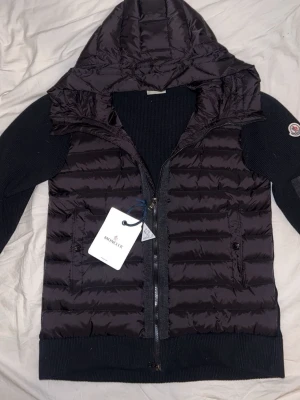 Svart dunjacka med huva från Moncler - Snygg svart dunjacka från Moncler med stickade ärmar och huva. Jackan har dragkedja framtill, två sidofickor med knappar och en Moncler-logga på ärmen. Perfekt för kyliga dagar och riktigt trendig look.