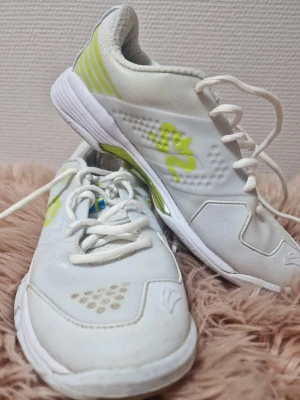 Vita sneakers med limegröna detaljer - Snygga vita sneakers med limegröna detaljer och snörning. Skorna har en sportig design med perforerade partier framtill och mönstrad sula. Tillverkade i syntetmaterial och textil, perfekta för en aktiv stil.
