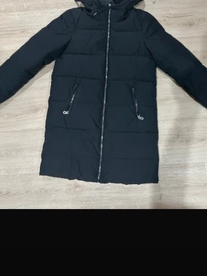 Svart lång dunjacka med huva - Snygg svart dunjacka med huva och långa ärmar. Jackan har två fickor med dragkedja och en rak, quiltad design. Perfekt för kalla dagar och har en stilren look som passar till det mesta.