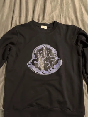Svart sweatshirt från Moncler - Säljer en svart sweatshirt från Moncler med ett stort, psykedeliskt tryck i lila och vitt på bröstet där det står 'Moncler'. Säljer denna eftersom den inte passar längre.