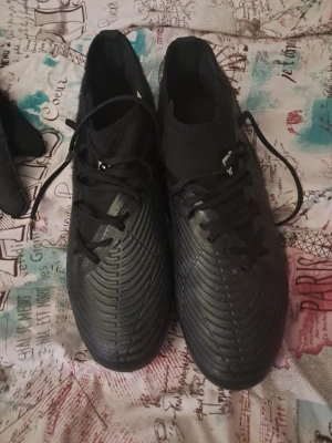 Svarta Adidas fotbollsskor med dobbar - Säljer ett par svarta Adidas fotbollsskor med blå dobbar och klassiska vita ränder på sidan. Skorna har snörning och är tillverkade i syntetmaterial med en modern, strömlinjeformad design. Perfekta för dig som vill ha stilrena och funktionella skor på planen.