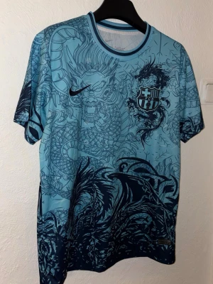 Blå FC Barcelona Nike tröja drakmönster - Snygg ljusblå FC Barcelona tröja från Nike med unikt drakmönster i mörkblått över hela plagget. Kortärmad modell med rund hals och klubbmärke på bröstet. Perfekt för dig som vill sticka ut på träningen eller matchen.