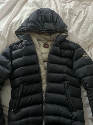 RESERVERAD!!! - Säljer en Mörkblå pufferjacka från Colmar med huva!. Jackan har en beige insida och diskret Colmar-logga på ärmen. Perfekt för kalla dagar och riktigt skön att bära. Materialet är syntet och jackan har en klassisk, quiltad look. Obs!!! Bild 3&4 visar hur dragkedjan till båda fickor endast funkat till halva!