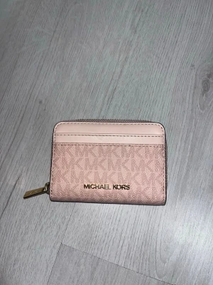 Ljusrosa plånbok från Michael Kors - Snygg ljusrosa äkta plånbok från Michael Kors med guldfärgad dragkedja och logga framtill. Plånboken har flera kortfack och ett större fack inuti. Tillverkad i tåligt konstläder med MK-mönster och stilren design. Aldrig använd!