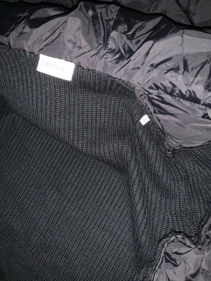 Svart cardigan från Moncler - Snygg svart cardigan från Moncler med stickad insida och glansig utsida. Cardigan har en modern look och är tillverkad i dunmaterial för extra värme. Perfekt för lager på lager och passar till många olika stilar.