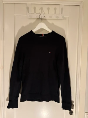Svart stickad tröja Tommy Hilfiger - Klassisk svart stickad tröja från Tommy Hilfiger i Pima cotton blend med cashmere. Tröjan har rund halsringning, diskret logga på bröstet och ribbade muddar vid ärmslut och nederkant. Perfekt för en stilren och enkel look.