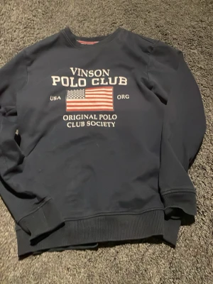 Svart sweatshirt Vinson Polo Club - Svart sweatshirt från Vinson Polo Club med tryck framtill i vitt och amerikansk flagga. Rund halsringning, ribbade muddar vid ärmslut och nederkant. Perfekt för en avslappnad och sportig stil.
