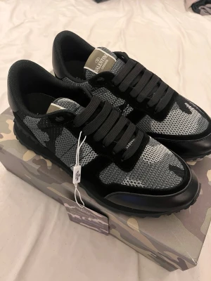 Valentino Garavani camo sneakers - Snygga Valentino Garavani sneakers i svart och grått med camouflagemönster. Skorna har snörning, meshpaneler och detaljer i skinn och mocka. Sitter på en platt sula med rund tå och coola taggade kanter.