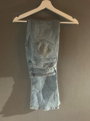 Ljusblå bootcut jeans med broderi och låg midja - Säljer ett par ljusblå bootcut jeans med snyggt broderat mönster på bakfickan. Jeansen har klassisk femficksdesign, dragkedja och knappstängning. Dom är lågmidjade. Perfekta för dig som gillar en avslappnad men ändå trendig look.