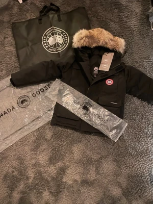 Svart Canada Goose Expedition dunjacka - Säljer en svart Canada Goose Expedition dunjacka med stor pälskant som går ta av på huvan och klassisk logga på bröstet. Jackan har flera fickor, dragkedja och kardborrestängning. Perfekt för kalla dagar och riktigt snygg vinterstil.