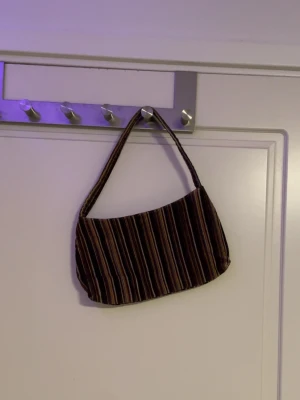 Randig brun handväska i tyg - Snygg handväska med bruna, svarta och beige ränder. Väskan har en mjuk, avlång form och ett kort axelband. Perfekt för dig som gillar retrovibbar och vill ha en unik accessoar till din outfit. Tillverkad i tygmaterial.