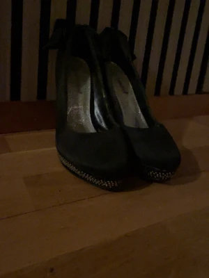 Svarta pumps med rosett och strass - Säljer ett par svarta pumps från Graceland i satinliknande material. Skorna har hög klack och plattform täckt med glittrande strassdetaljer. Baktill sitter en stor rosett som ger extra statement. Perfekta för dig som vill sticka ut! Även billigare vid snabb affär!