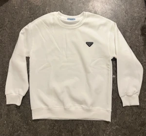 Vit sweatshirt från Prada - Säljer en clean vit sweatshirt från Prada med det ikoniska svarta trekantsloggan på bröstet. Tröjan har rund halsringning, ribbade muddar och är gjord i mjukt material som känns skönt mot huden. Perfekt för dig som gillar minimalistisk och lyxig stil.