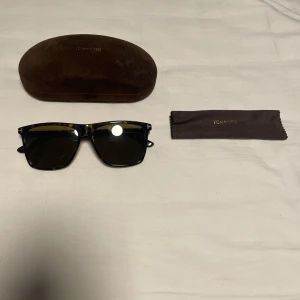 Svarta solglasögon Tom Ford - Snygga svarta solglasögon från Tom Ford med klassisk rektangulär form och mörka linser. Bågarna har subtilt mönster i mörkbrun och svart, och loggan syns på skalmen. UV400-skydd och medföljande fodral och putsduk ingår.