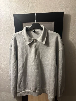 Grå halvzip sweatshirt med krage - En stilren grå sweatshirt med halv dragkedja och bred krage. Tröjan har en avslappnad passform, långa ärmar och ribbade muddar vid ärmslut och nederkant. Perfekt för en casual look och enkel att matcha med jeans eller mjukisbyxor.
