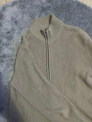 Beige ribbad stickad kofta med dragkedja - Säljer en beige stickad kofta med ribbad struktur och hög krage. Koftan har en hel dragkedja framtill och långa ärmar. Perfekt för lager på lager under kyligare dagar. Enkel och stilren design som passar till det mesta.