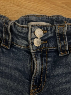 Blå bootcut jeans från Gina Tricot - Säljer ett par klassiska blå jeans från Gina Tricot i storlek 158. Jeansen har bootcut-modell, två knappar i midjan och snygga slitningar framtill. Materialet är jeans med kontrastsömmar och femficksdesign. 