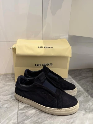  Axel Arigato skor - Säljer ett par navy blue sneakers från Axel Arigato i mocka med vit sula och minimalistisk design. Skorna har en låg profil och diskret logga på sidan. Perfekta för dig som gillar stilrena och moderna skor. Box och dustbag medföljer