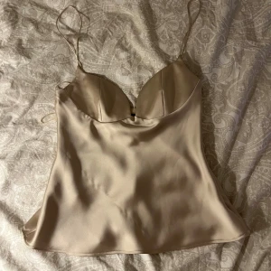 Satin topp Zara - Säljer en champagnefärgad linnetopp i satin med tunna axelband och lätt formade kupor. Toppen har en slät, glansig yta och en enkel, elegant siluett som ger en lyxig känsla. Perfekt att styla med jeans eller kjol för en trendig look. Lite slitning på sista bild