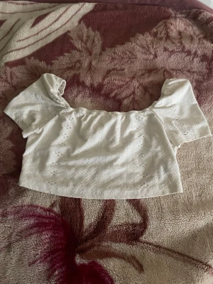 Vit croppad offshoulder blus - Söt vit croppad blus med offshoulder-design och korta puffärmar. Blusen har broderade detaljer och är tillverkad i ett lätt bomullstyg som passar perfekt till varma dagar. Perfekt att styla med höga jeans eller kjol.