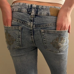 Bootcut ljusblå jeans med broderi - Säljer ett par ljusblå bootcut jeans med snyggt guldbroderi på bakfickorna. Jeansen har låg midja och klassisk femficksdesign. Materialet är jeans i bomull och passformen är normal. Perfekta för dig som gillar detaljer och en retrovibe.