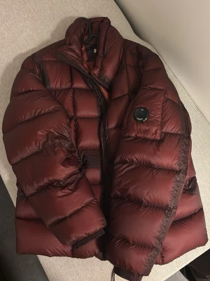 Vinröd pufferjacka från C.P. Company - Säljer en vinröd pufferjacka från C.P. Company med ikonisk linsdetalj på ärmen. Jackan är dunfodrad och har en quiltad design med dragkedja framtill. Perfekt för kalla dagar och har en riktigt schysst streetstil.