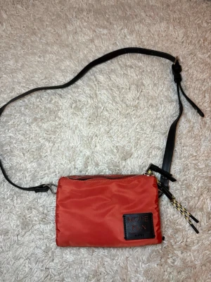 Orange axelväska från Parfois - Säljer en orange axelväska från Parfois med svart justerbart axelband och två separata fack med dragkedja. Väskan har en svart patch med texten 'MORE FUN' framtill och coola detaljer i snöre. Perfekt för dig som vill ha en smidig och snygg väska till vardags.                                                             Väskan är använd en gång annars är den helt ny. 