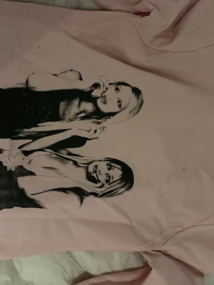 Ljusrosa hoodie med tryck - Säljer en ljusrosa hoodie med dragkedja och fickor framtill. På ryggen finns ett svartvitt tryck med två tjejer i retrostil. Hoodien har huva och är gjord i mjukt bomullsmaterial. Perfekt för dig som gillar streetwear och vill sticka ut lite extra.