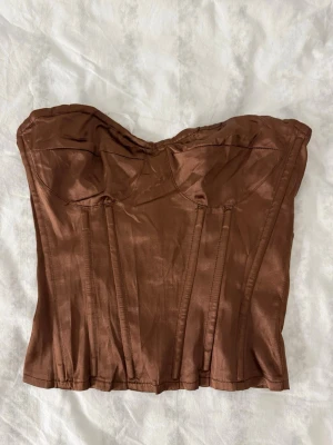 Brun satin korsett med bygel - Snygg brun korsett i glansigt satinmaterial med bygel och markerade sömmar framtill. Axelbandslös design.