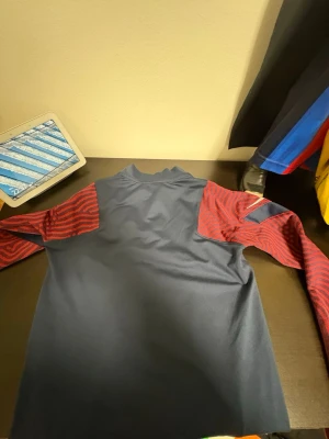 PSG träningströja blå/röd Nike - Säljer en Paris Saint-Germain träningströja från Nike i marinblått med röda, mönstrade ärmar och vita detaljer. Tröjan har halv dragkedja, PSG-logga på bröstet och sponsortryck framtill. Perfekt för fotbollsträning eller som supporterplagg.