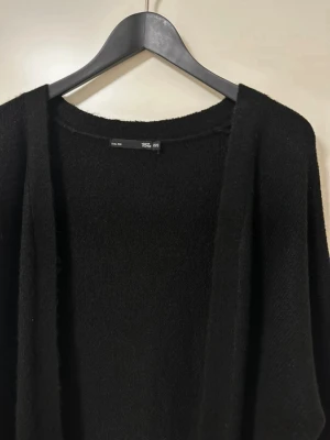 Svart lång kofta från 157 - Mysig svart lång kofta från 157 med öppen front och två fickor framtill. Koftan är stickad och har långa ärmar samt ribbade muddar vid ärmslut och nederkant. Perfekt att slänga över en t-shirt eller hoodie för en chill look.