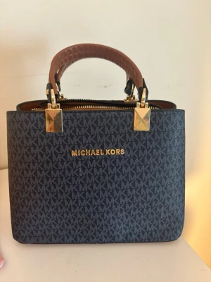  handväska  Michael Kors liknande  - Mörkblå  handväska  Michael Kors liknande med guldiga detaljer och klassiskt MK-mönster över hela väskan. Handtaget är brunt och väskan har dragkedja upptill. Insidan är fodrad med logotypmönster och har flera fack för smart organisering.