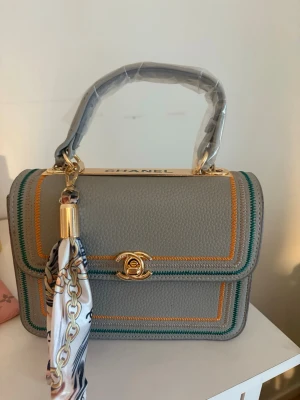 Grå handväska  med scarf - Säljer en grå handväska  i skinn med gulddetaljer och färgglada sömmar i orange och grönt. Väskan har ett elegant handtag och en snygg scarf med kedjemönster som accessoar. Klassisk twist-lock framtill och rymligt innerfack. Perfekt för dig som vill sticka ut.