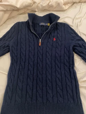 Mörkblå kabelstickad tröja Polo Ralph Lauren - Mörkblå kabelstickad tröja från Polo Ralph Lauren med halv dragkedja och hög krage. Tröjan har ett rött broderat logomärke på bröstet och ribbade muddar vid ärmslut och nederkant. Aldrig använd, de är storlek M men passar perfekt som S. Kom med frågor o prisförslag