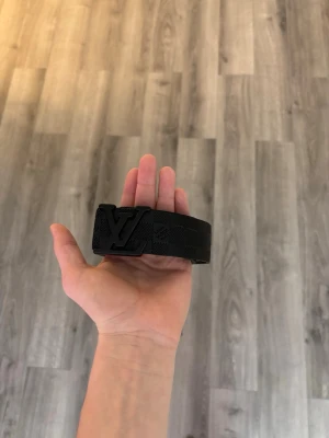 Svart bälte - Snyggt svart bälte  med klassiskt rutmönster och stor logga på metallspännet. Bältet är gjort i ett tåligt material och har en modern, stilren look som passar till jeans eller byxor. Tar emit rimliga bud.