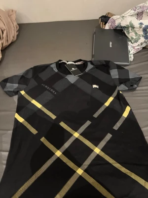 Burberry rutig t-shirt S-M - Snygg t-shirt från Burberry med klassiskt rutigt mönster i svart, grått och gult. T-shirten har rund halsringning och korta ärmar. Perfekt för dig som vill sticka ut med en ikonisk design och exklusiv känsla.